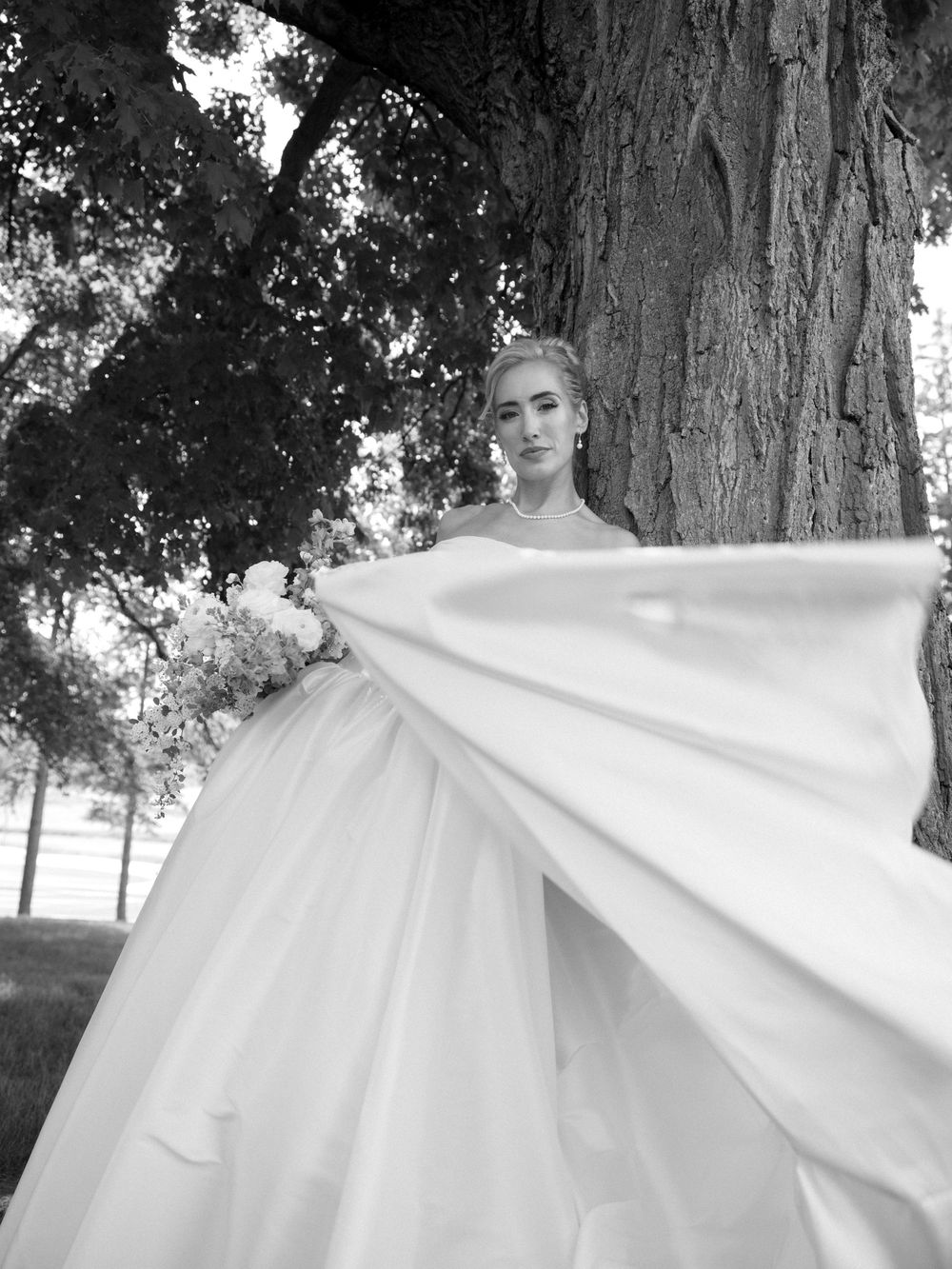 Bride in a stunning ballgown twirling her cape beside a large tree in a black and white photo.