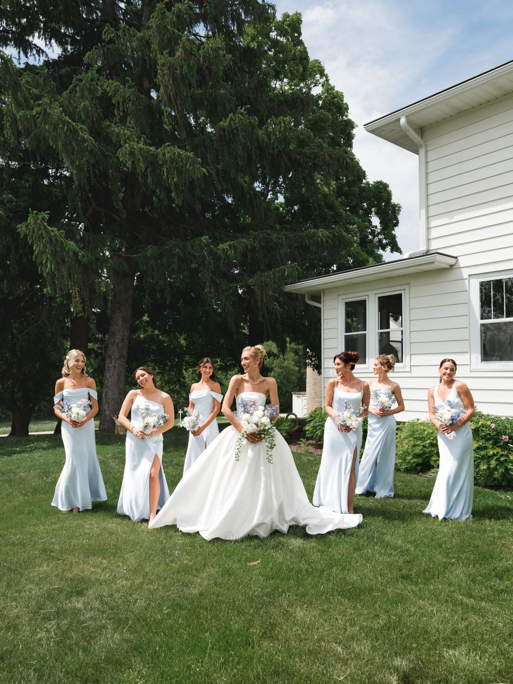 Bride in white ball gown poses with six bridesmaids in light blue strapless gowns on green lawn beside white house.