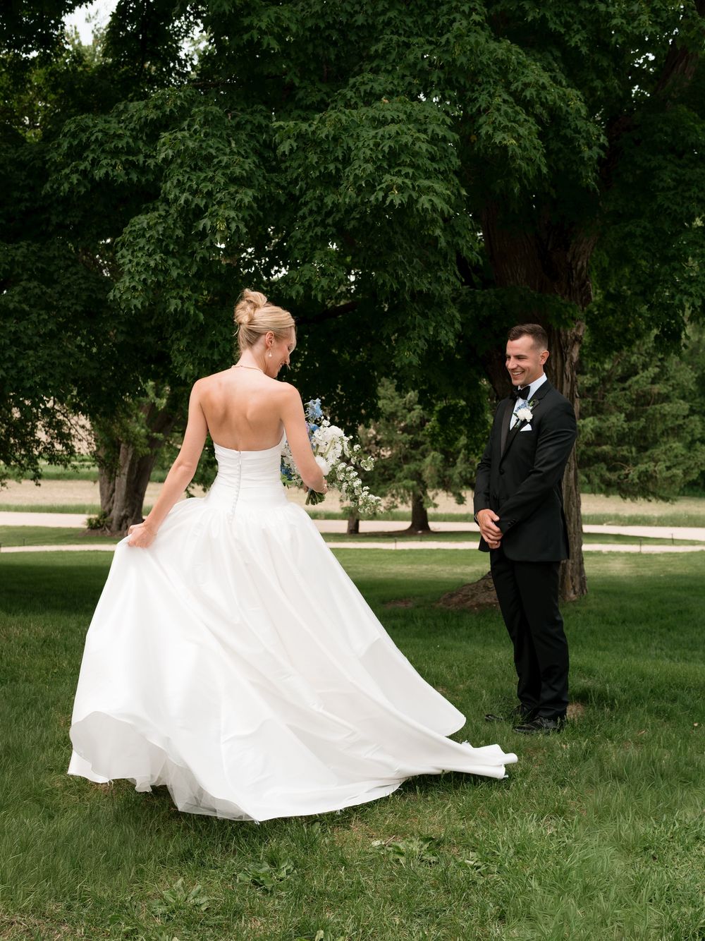 Bride in strapless white ball gown approaches smiling groom during outdoor first look ceremony.