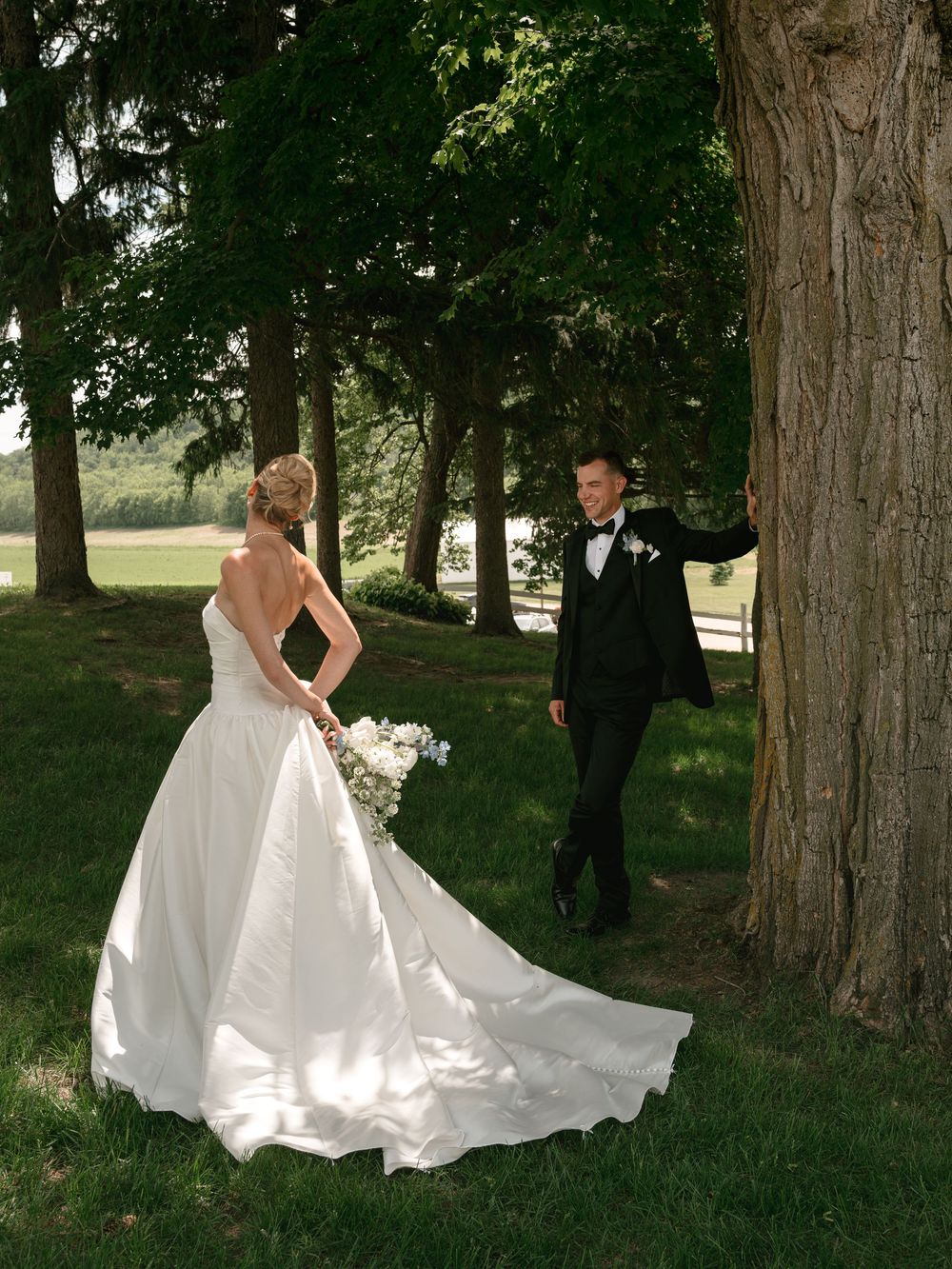 Bride in white gown with train faces groom in black tuxedo leaning against large tree outdoors on wedding day.