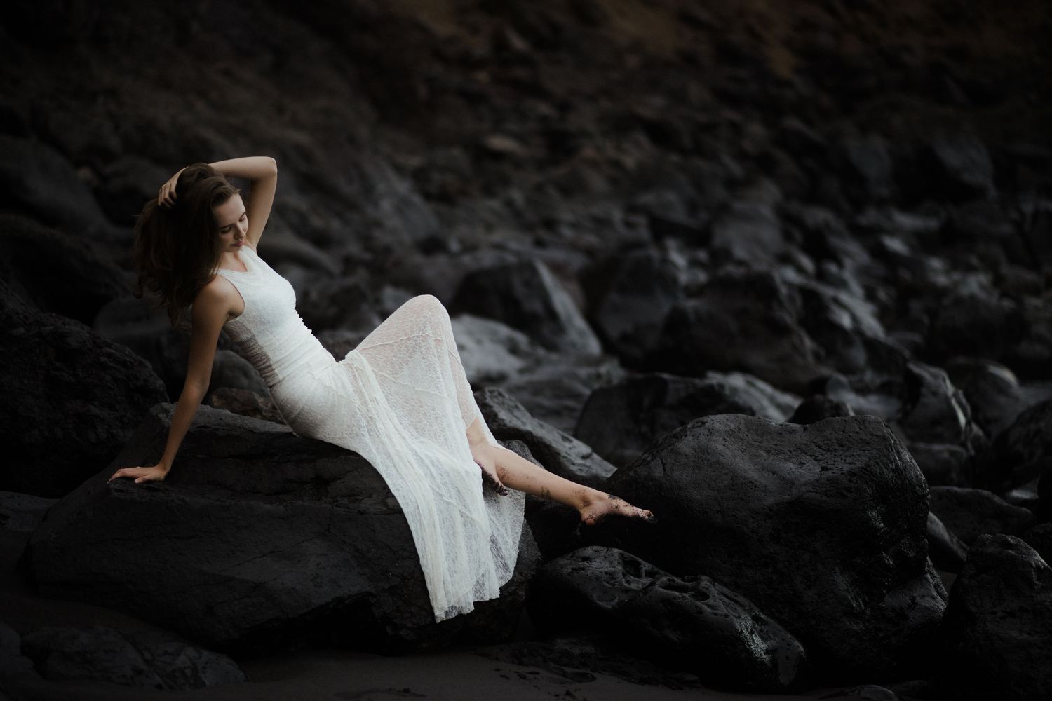 Woman in white dress with pearl earrings gazes upward against rocky backdrop.
