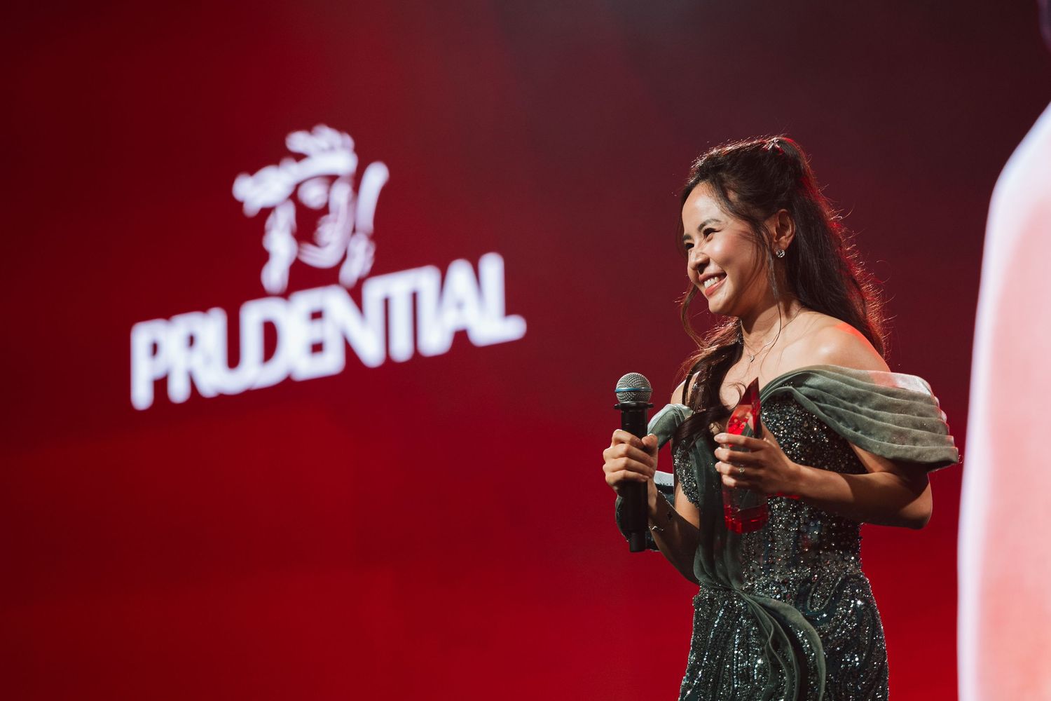 Woman in green dress speaks on stage at Prudential Agency Award 2026 event, holding microphone under red lighting.