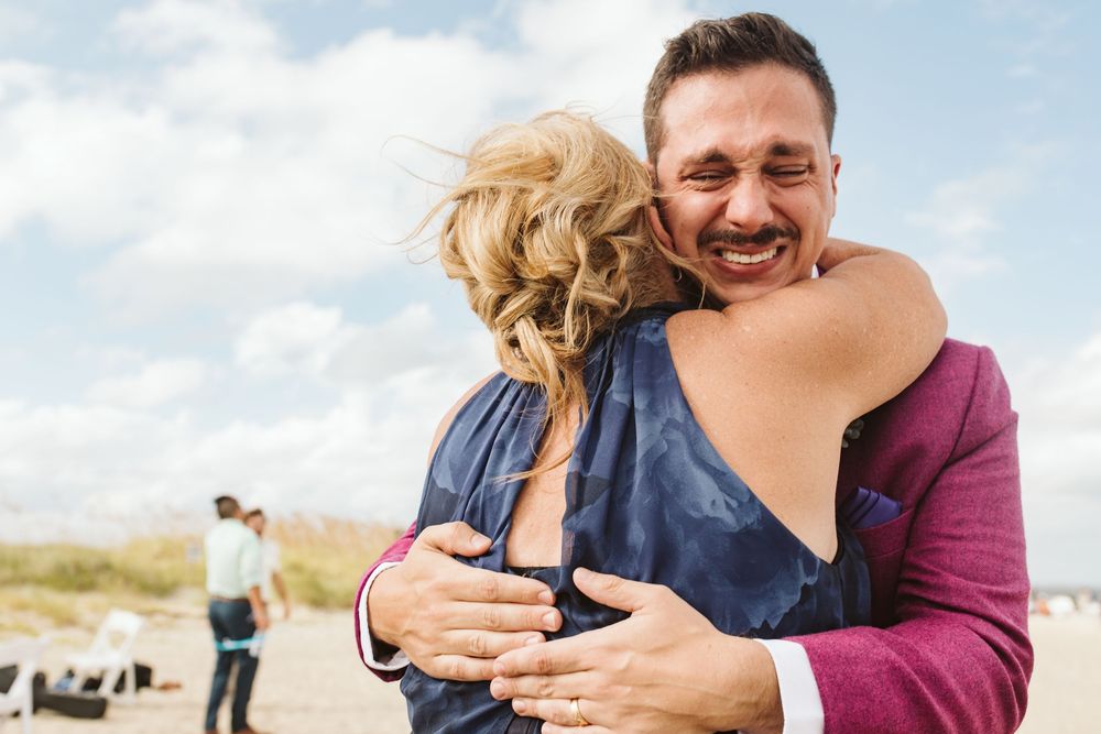 A man in a purple suit smiles while hugging a woman in a blue dress on a beach under a cloudy sky.