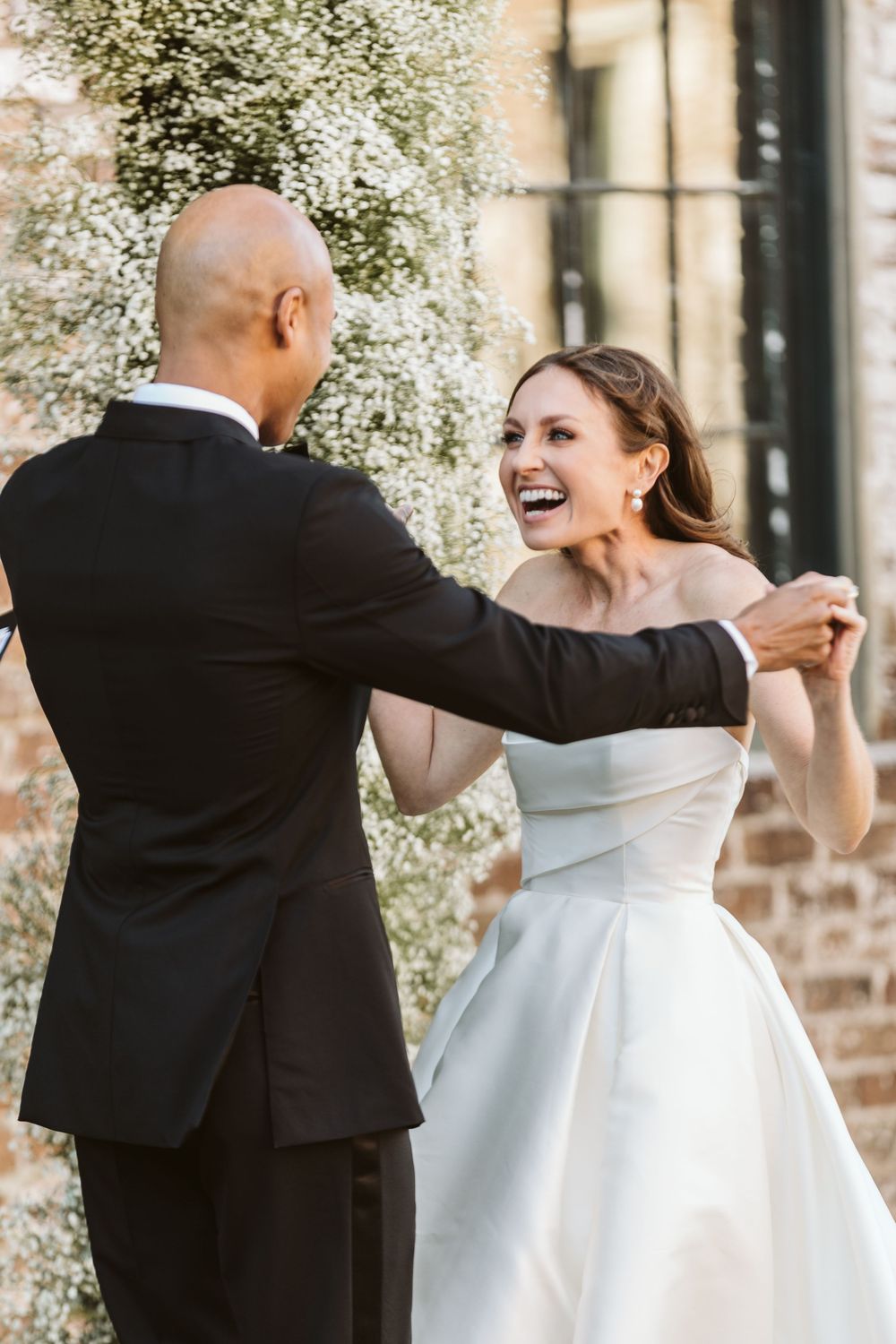 Bride in off-shoulder white gown laughs joyfully while dancing with groom in black tuxedo outdoors.