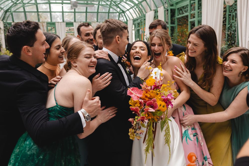 Bride and groom surrounded by joyful wedding party guests in a greenhouse venue, bride holding vibrant tropical bouquet.