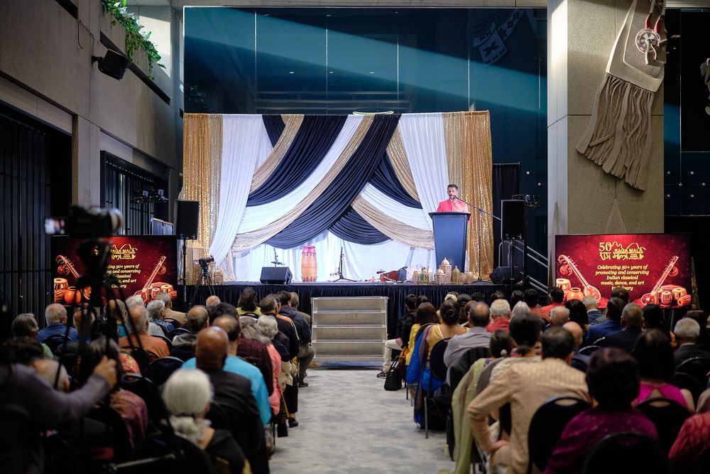 Large audience attends a formal event with a decorated stage, draped curtains, and a speaker at a podium.
