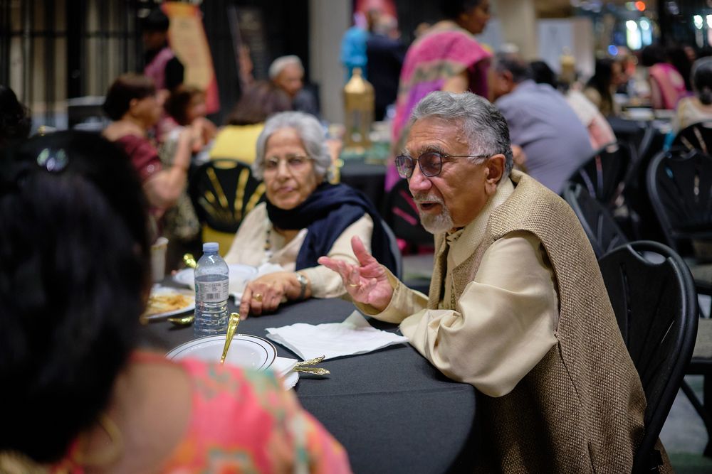 Elderly Indian man gestures while conversing at a social gathering dinner event with women seated nearby.