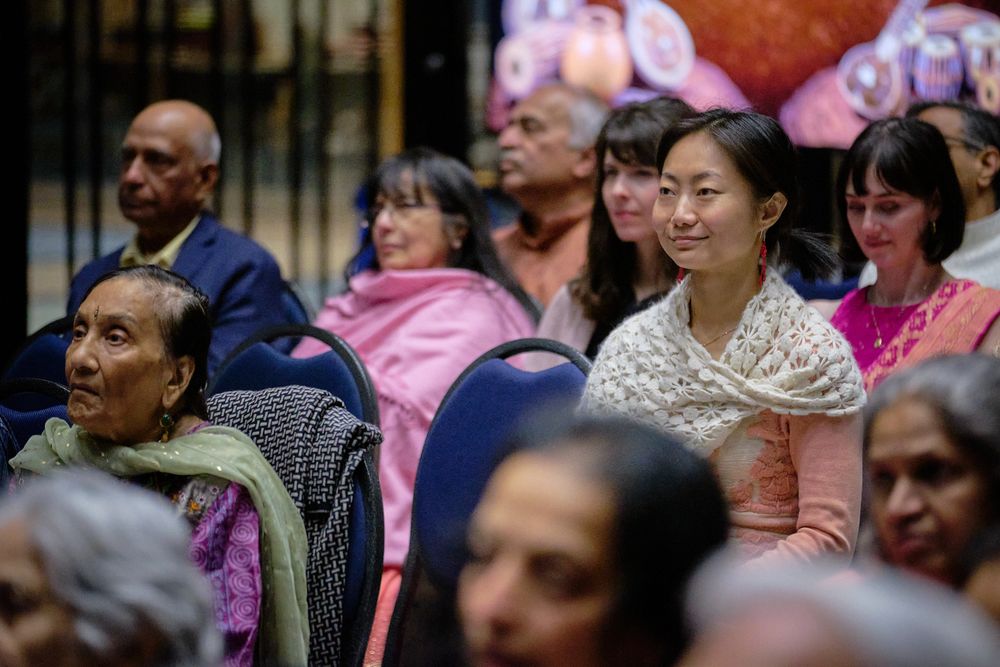 Diverse audience seated at a cultural event, smiling woman in foreground wearing traditional attire with pink balloons behind.