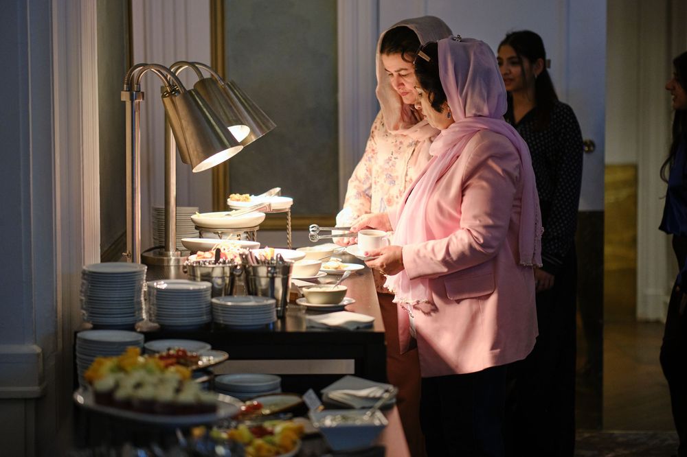 Two women serving themselves at an elegant buffet table with warm lamp lighting and stacked plates.