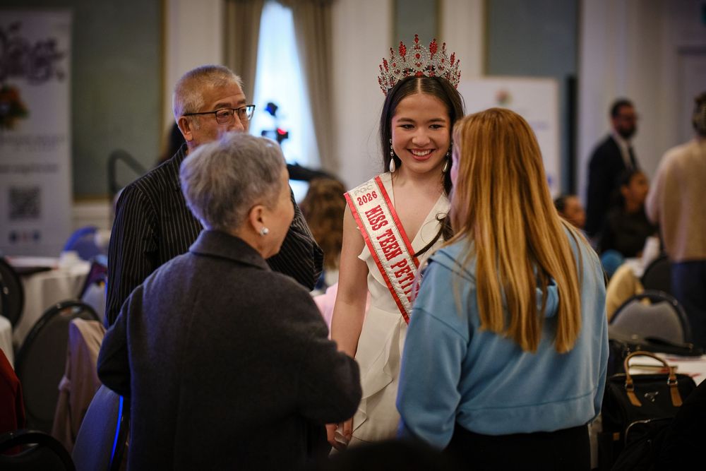 Young beauty queen in white gown and red crown smiles while greeting guests at a formal indoor event.
