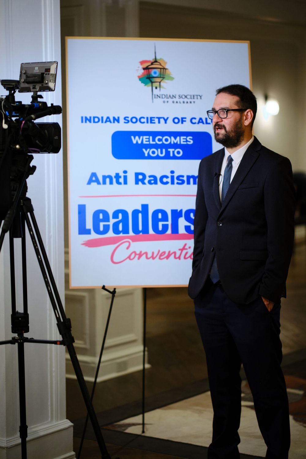 Man in suit stands beside Indian Society of Calgary Anti Racism Leaders Convention sign at formal event.