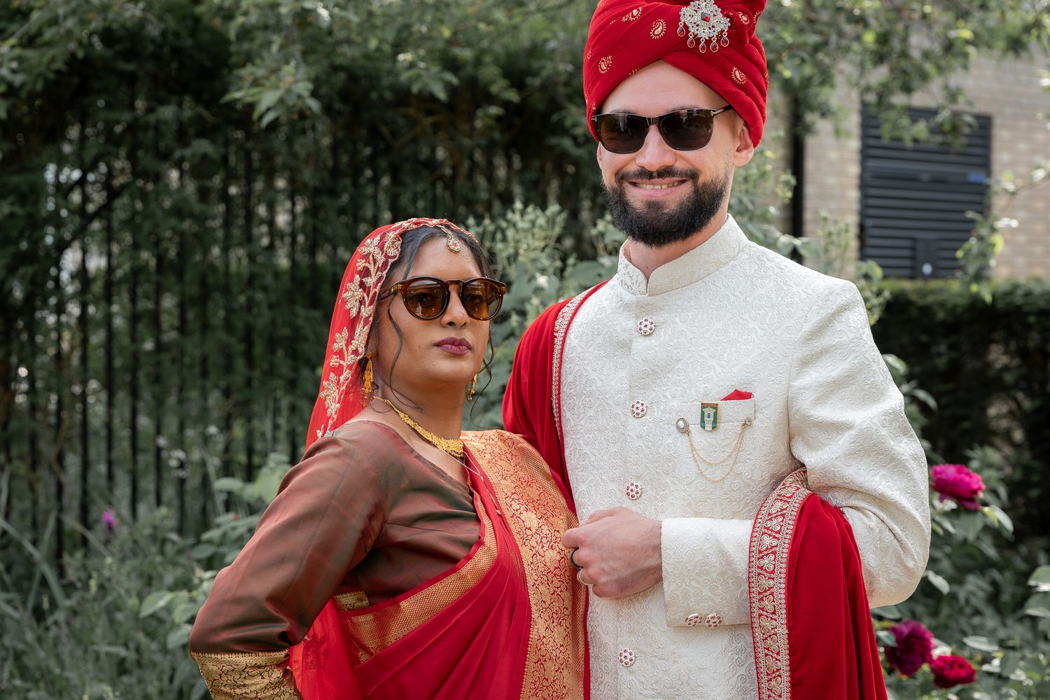 Couple in traditional Indian wedding attire, woman in red saree and man in white sherwani with red turban, posing outdoors.