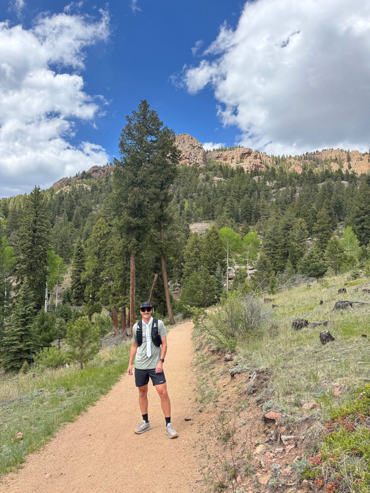 Hiker standing on a dirt trail surrounded by tall pine trees and rocky mountains under a blue sky.