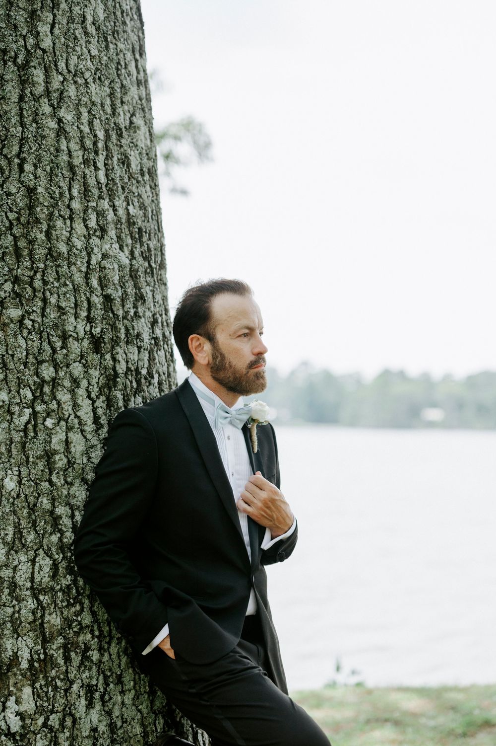 Bearded groom in black tuxedo with white bow tie leans against tree by lake, holding boutonniere.