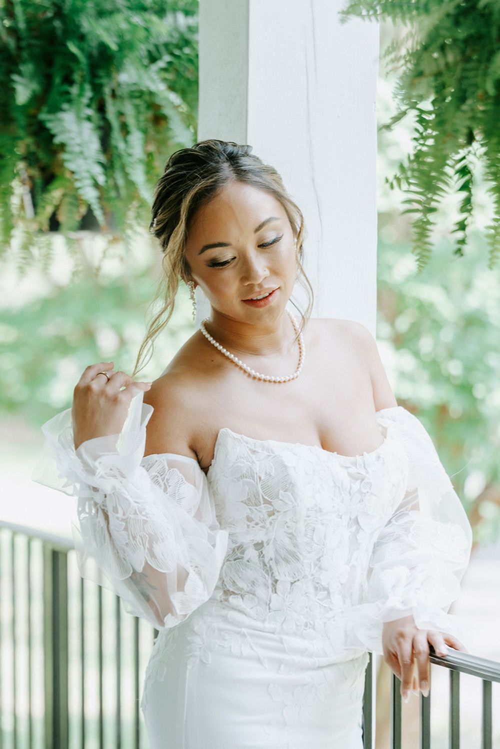 Beautiful bride in off-shoulder lace wedding gown with pearl necklace poses on a sunlit porch.