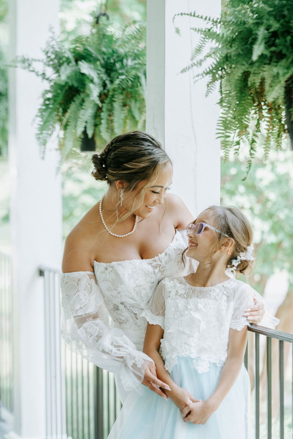 Bride in lace off-shoulder gown smiles with flower girl on a white porch surrounded by ferns.