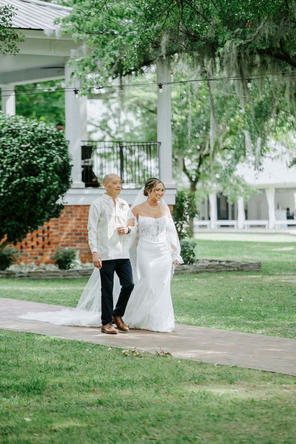 Bride in fitted lace gown walks with escort across green lawn toward white plantation-style venue.