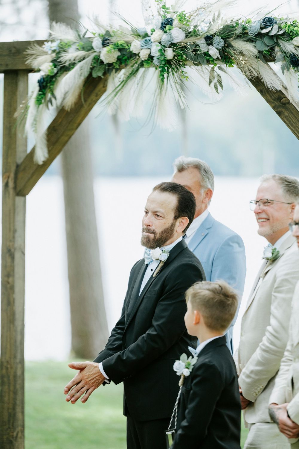 Emotional groom awaits his bride at a floral wooden arch during an outdoor lakeside wedding ceremony.