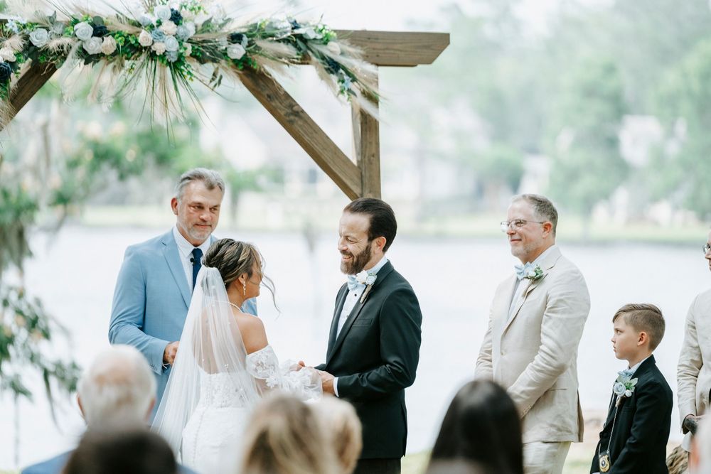 Outdoor wedding ceremony by a lake with bride and groom exchanging vows under a floral arch.