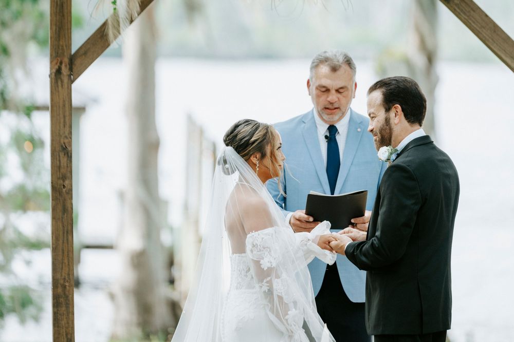Bride and groom exchange vows with officiant at outdoor waterfront wedding ceremony under wooden arch.