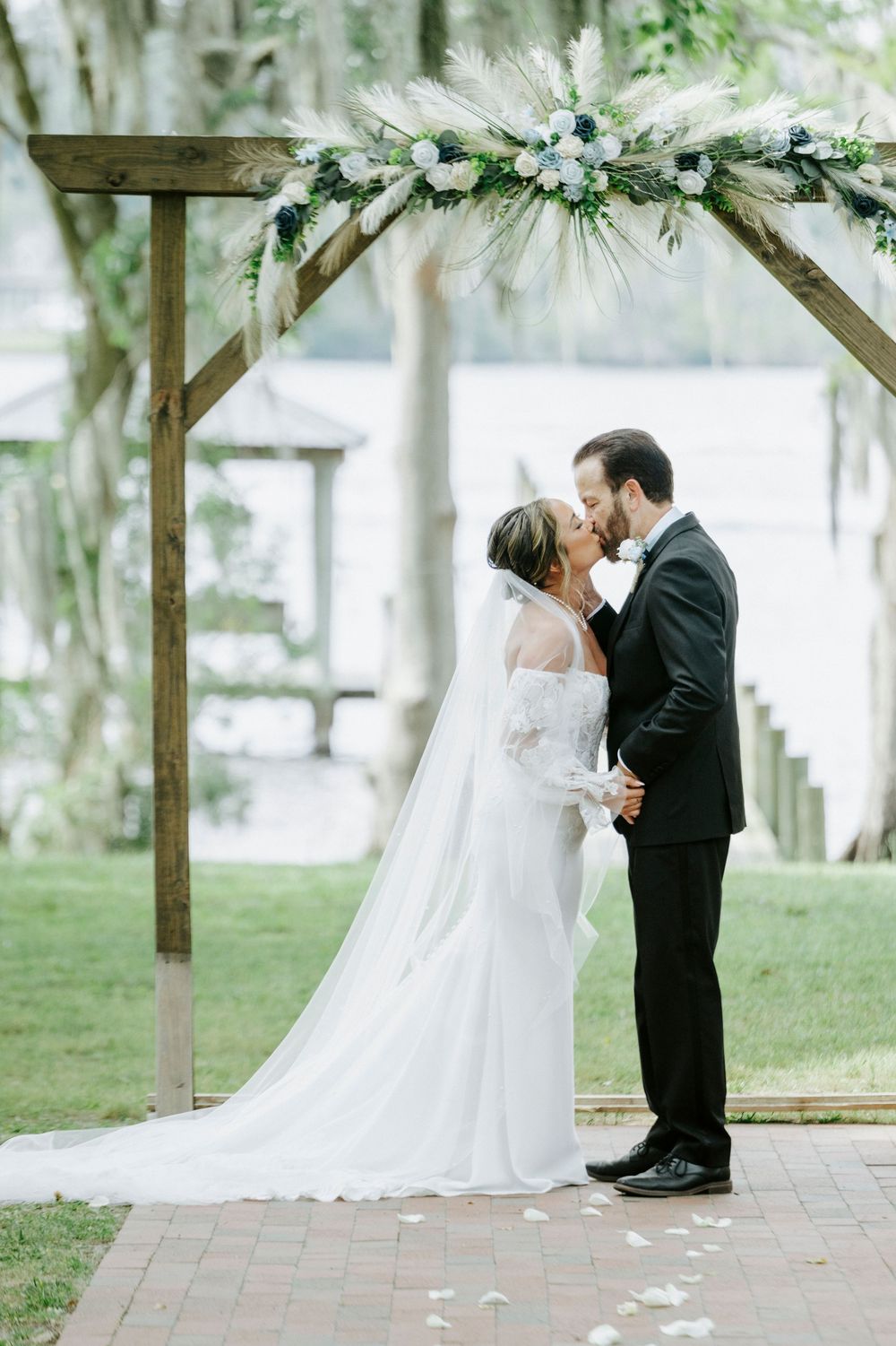 Bride and groom share first kiss under rustic wooden arch adorned with white floral arrangements.