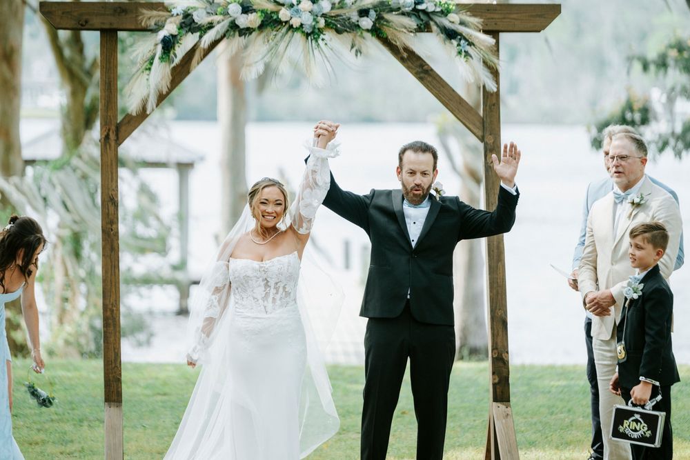 Bride and groom celebrating under a floral arch at an outdoor waterfront wedding ceremony with raised hands.