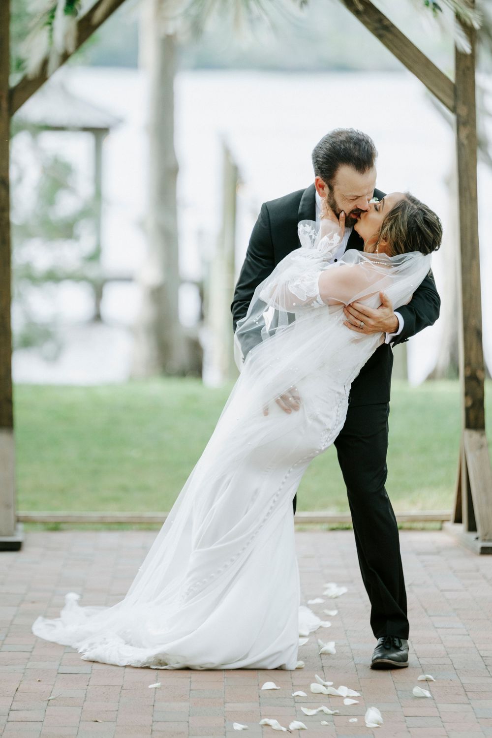 Groom dips bride for a romantic kiss under a wooden arch with rose petals on brick pathway at waterfront wedding.