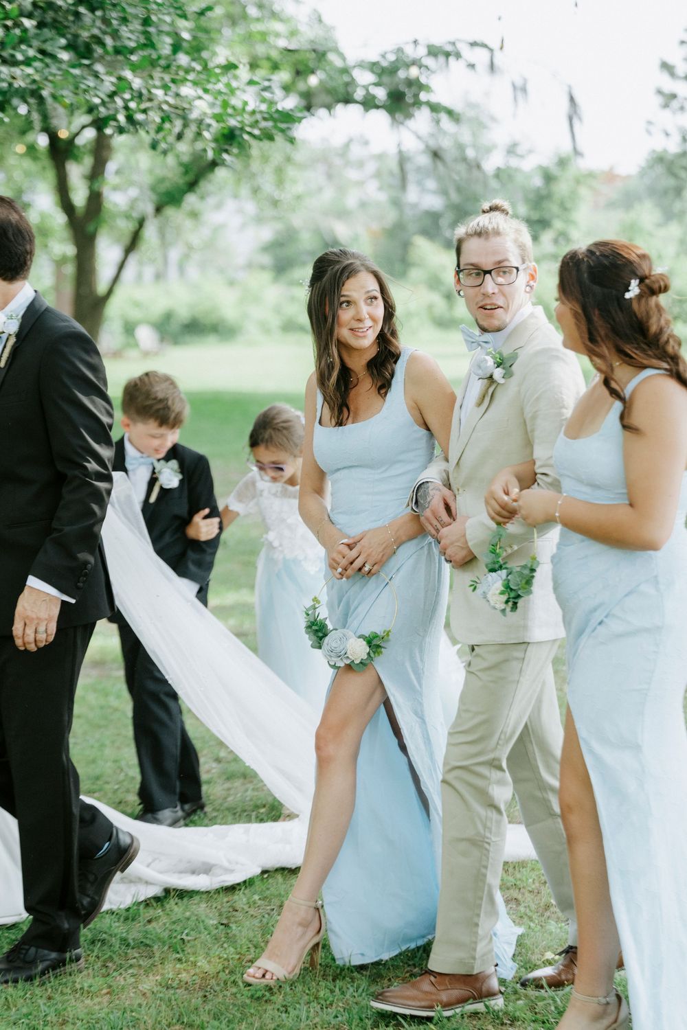 Bridesmaids in light blue dresses and groomsmen in tan suits gather outdoors at a summer wedding celebration.