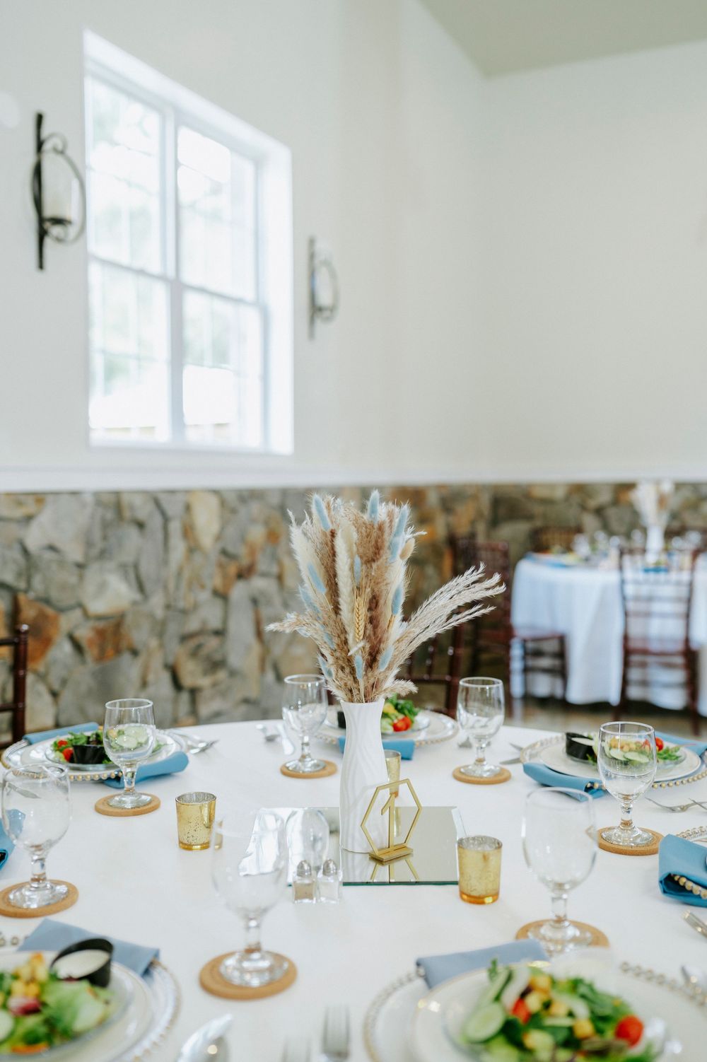 Elegant wedding reception table setting with pampas grass centerpiece, blue napkins, and gold accents.