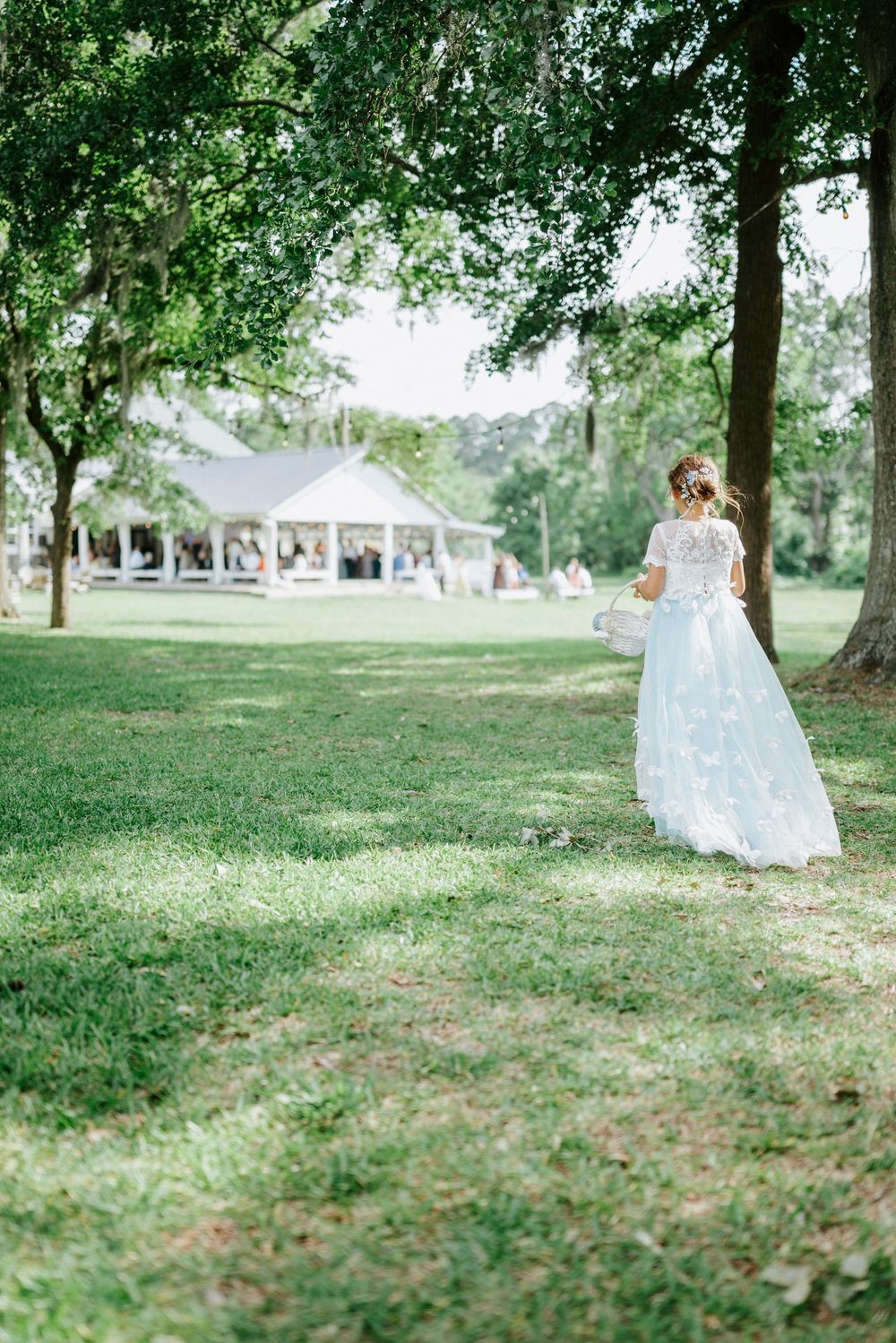 Bride in lace wedding gown walks across green lawn toward white pavilion surrounded by trees.