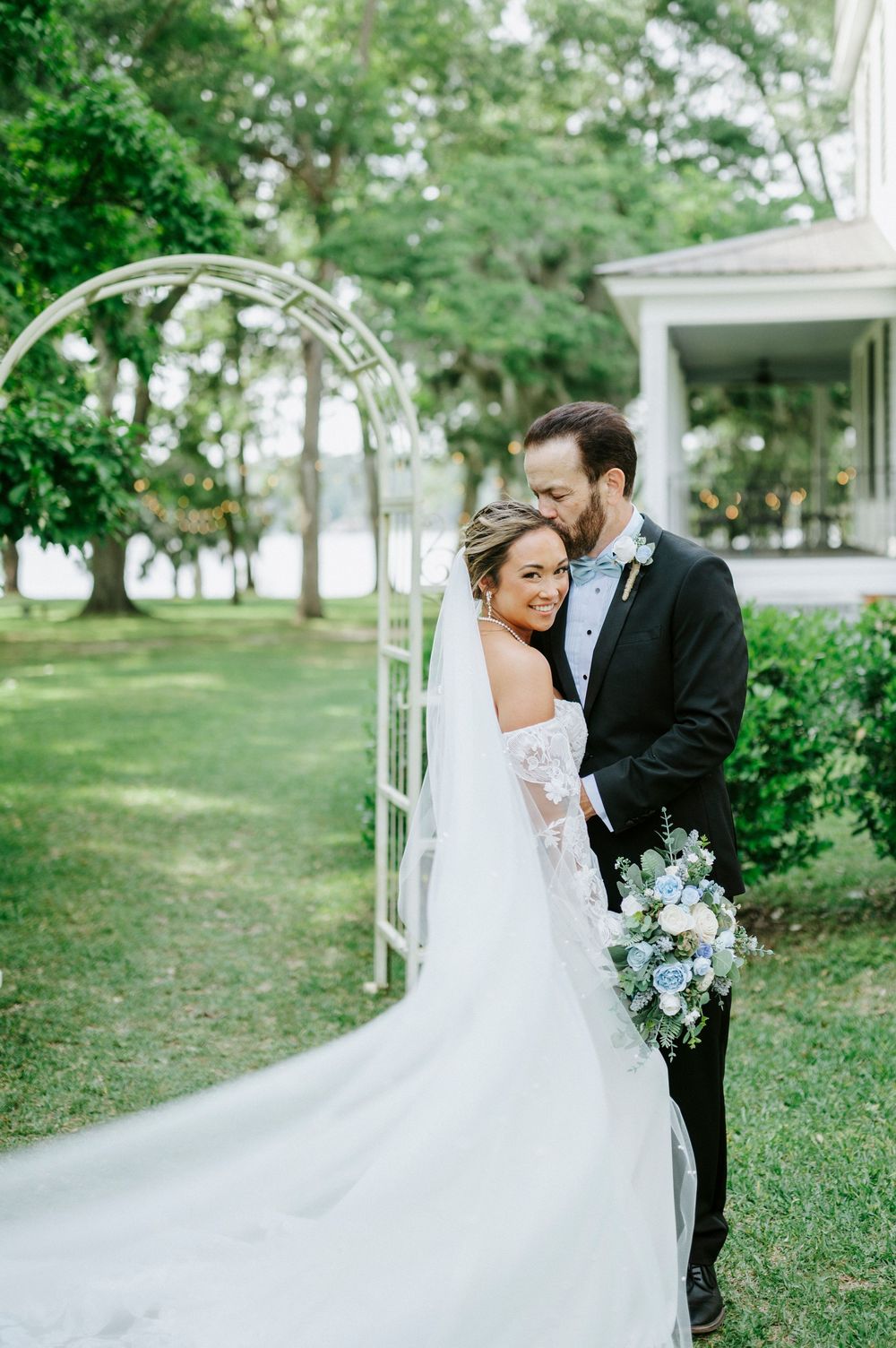 Bride and groom embrace under white garden arch, bride holding blue and white bouquet.
