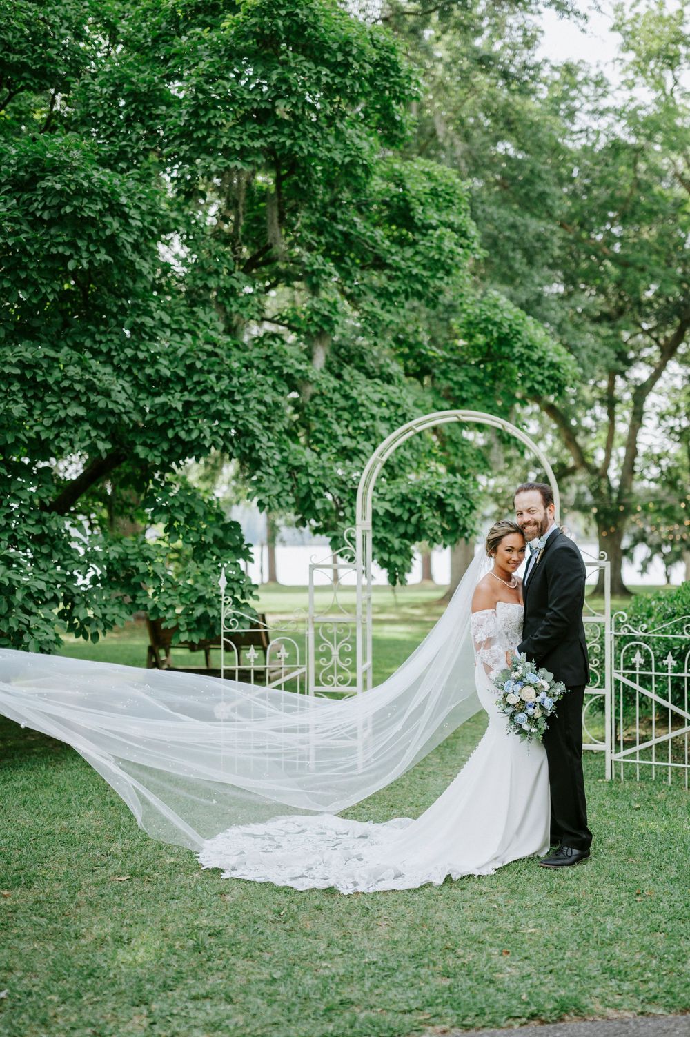 Bride in fitted white gown with flowing cathedral veil poses with tuxedo-clad groom in lush garden.