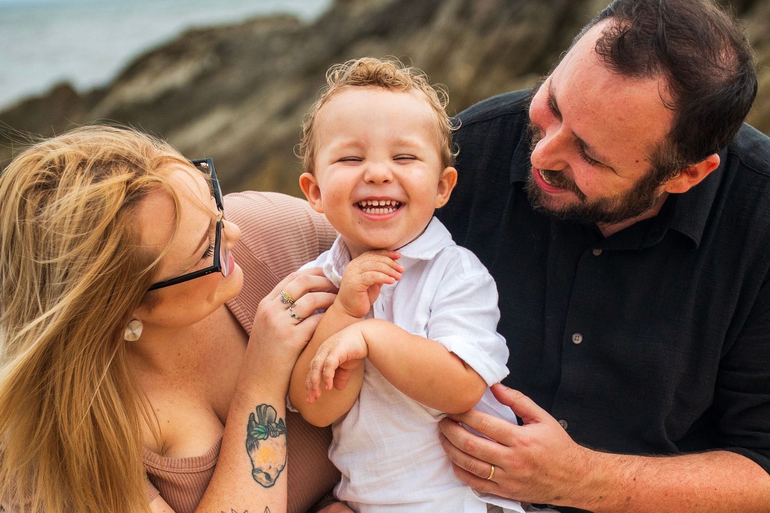 Happy toddler boy laughing with parents on rocky beach, family portrait outdoors.