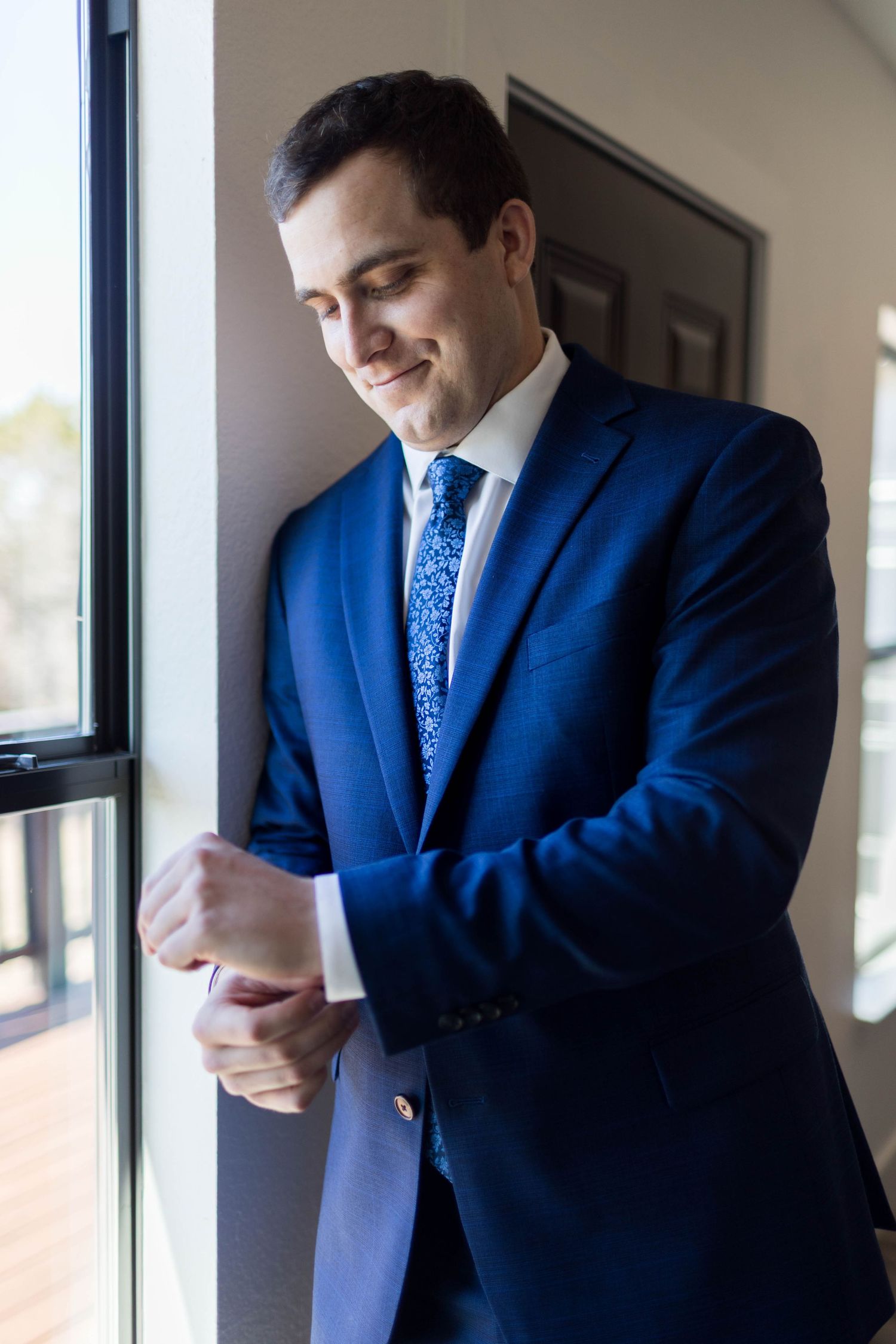 A smiling groom in a navy blue suit adjusts his cufflinks while standing by a window on his wedding day.