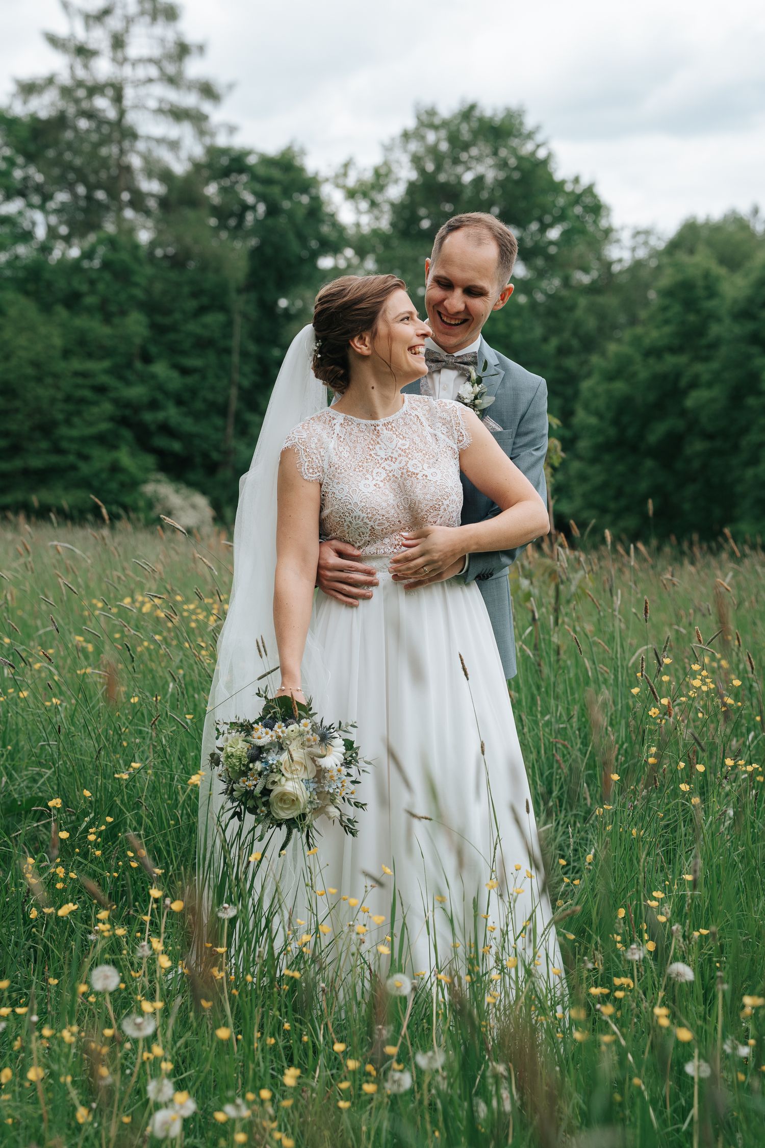 A joyful bride and groom embrace in a wildflower meadow, she holds a white bouquet, wearing a lace gown with veil.