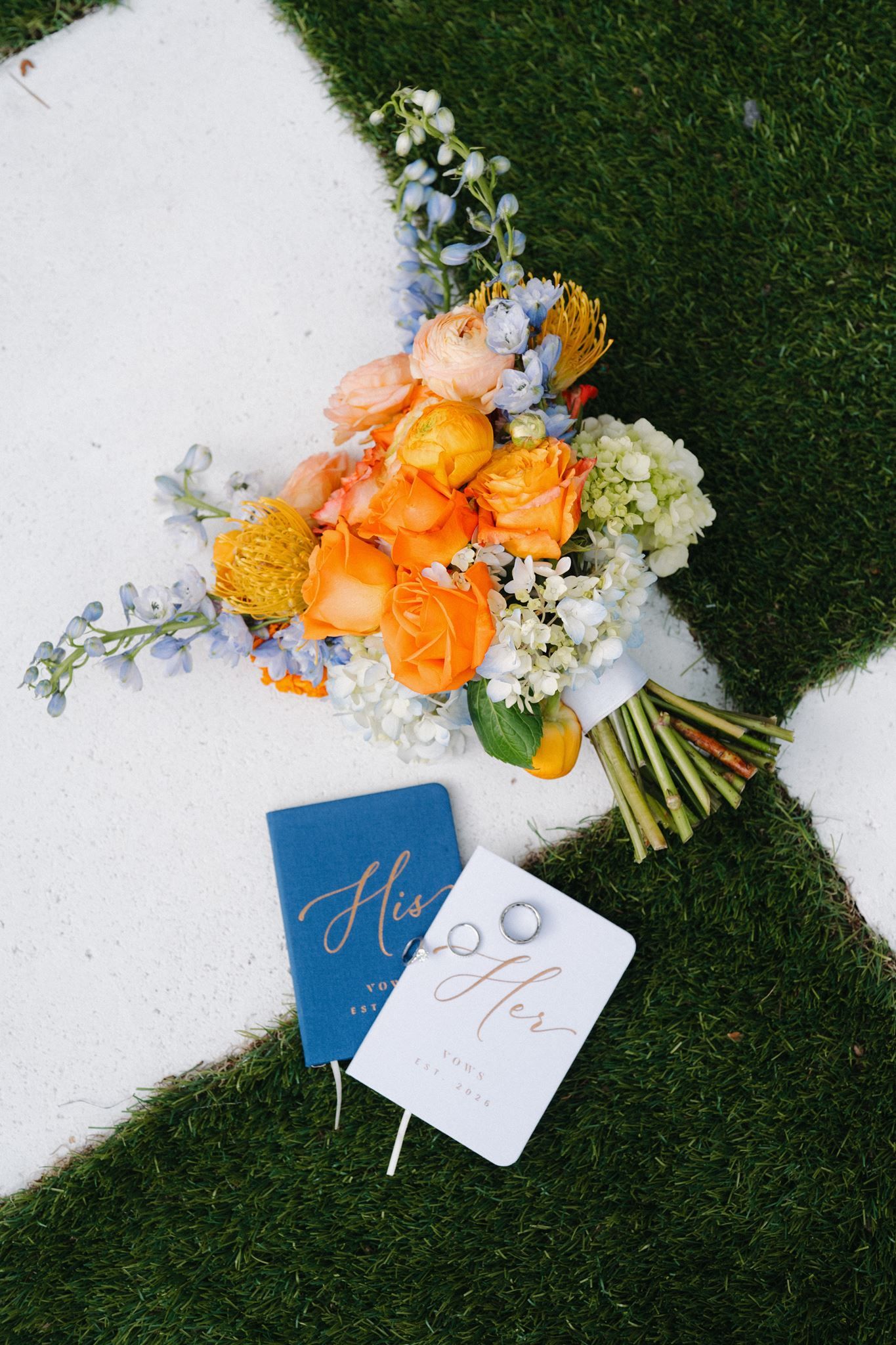 Wedding flat lay with orange bouquet, blue His & Hers vow books, and gold rings on green grass.