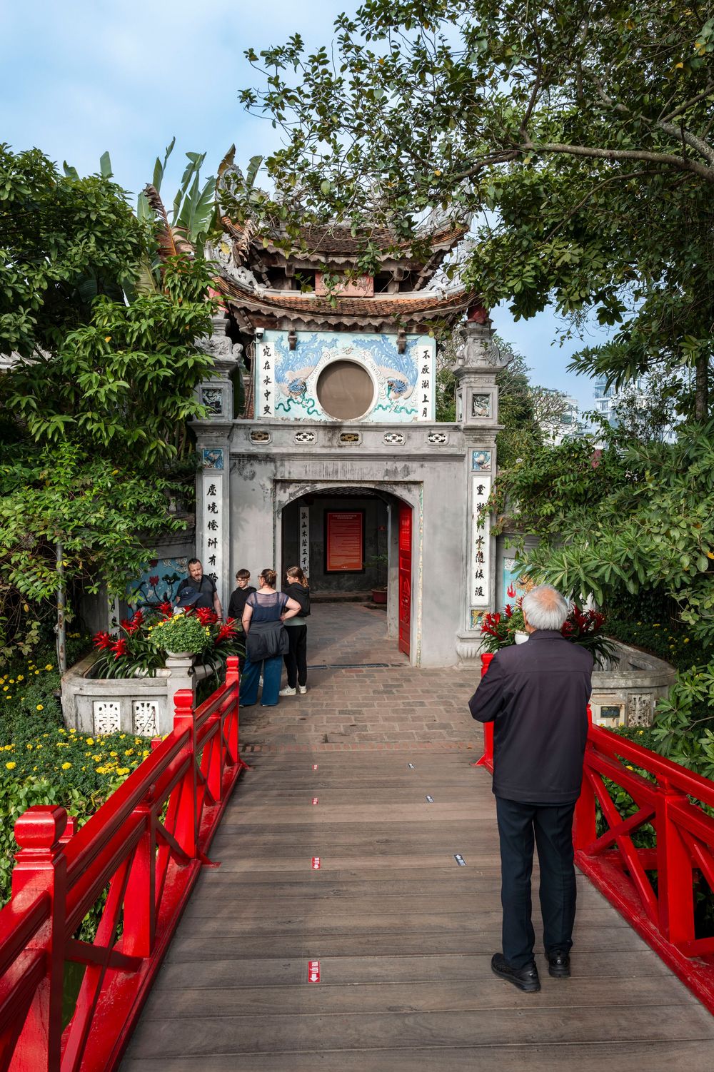 Red bridge leading to Ngoc Son Temple gate surrounded by lush trees at Hoan Kiem Lake, Hanoi, Vietnam.