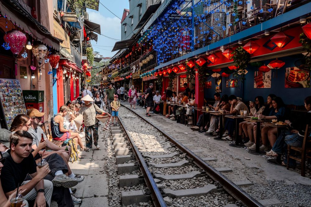 Tourists sitting along Hanoi Train Street with red lanterns decorating colorful cafes beside active railway tracks in Vietnam.