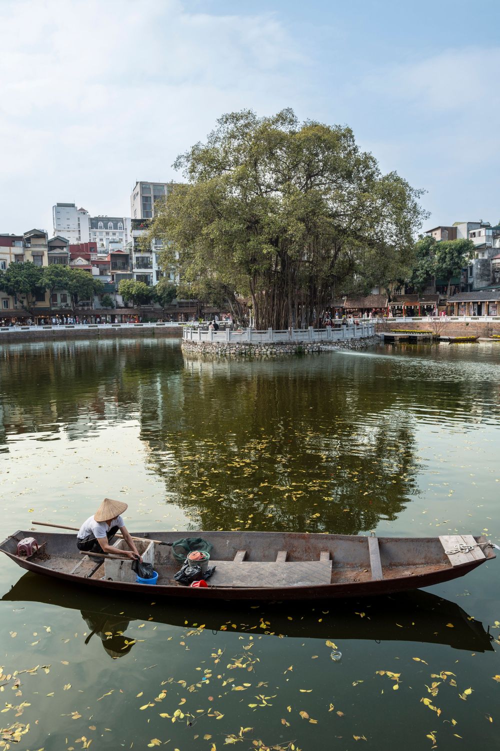 Local Vietnamese fisherman rowing wooden boat on Hoan Kiem Lake with historic temple island in background, Hanoi.