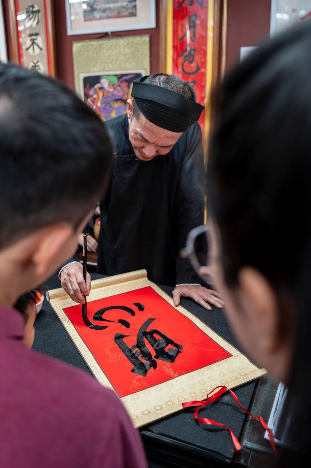 Traditional Chinese calligrapher in black robe writing on red paper scroll with brush, observed by students.