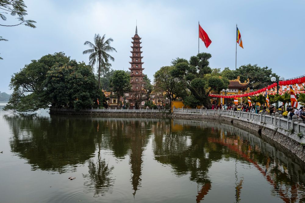 Tran Quoc Pagoda reflected in West Lake, Hanoi, Vietnam, with Vietnamese flags and lush trees surrounding the ancient tower.
