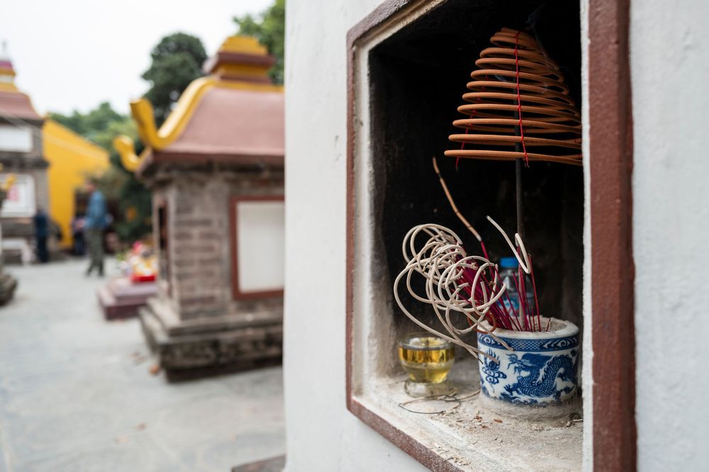 Spiral incense coils and blue ceramic pot in a temple niche, with colorful Buddhist shrine buildings in background.