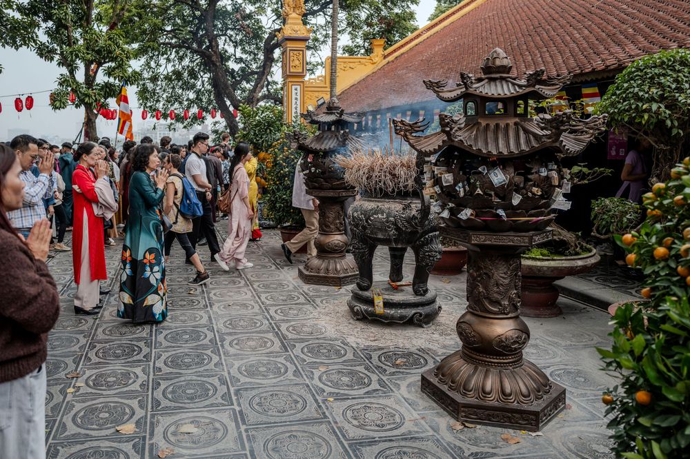 Visitors gather around ornate bronze incense burners at a traditional Vietnamese temple courtyard decorated with lanterns.