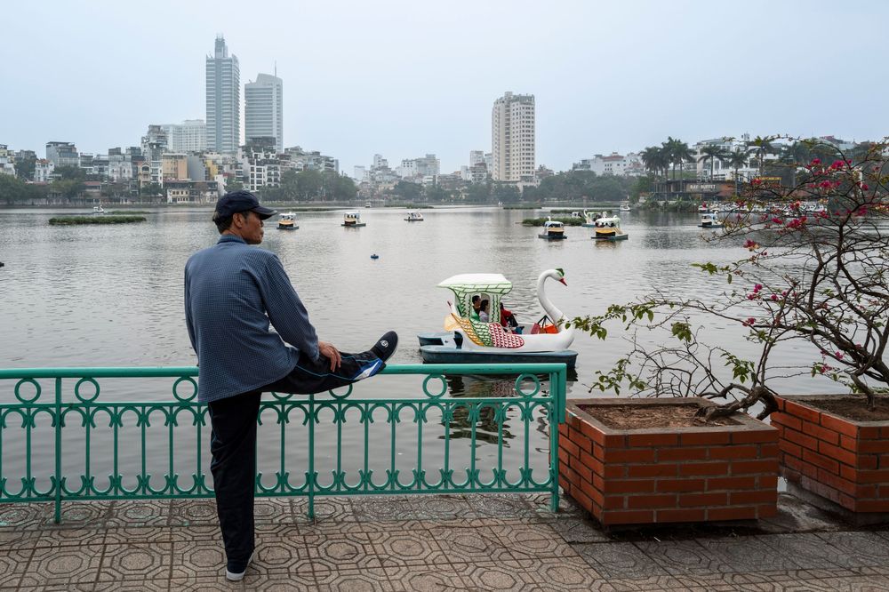 Man stretching by ornate green railing overlooking a busy harbor with boats and city skyline in overcast weather.