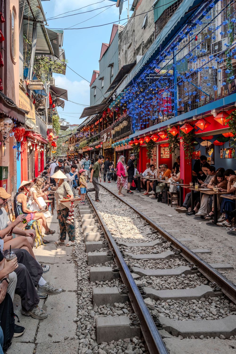 Tourists sit alongside active railway tracks lined with colorful red lanterns and cafes in Hanoi Train Street, Vietnam.