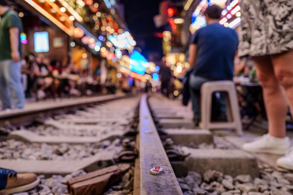 Night scene at Hanoi Train Street with bokeh lights, railway tracks, and tourists sitting along the narrow alley.