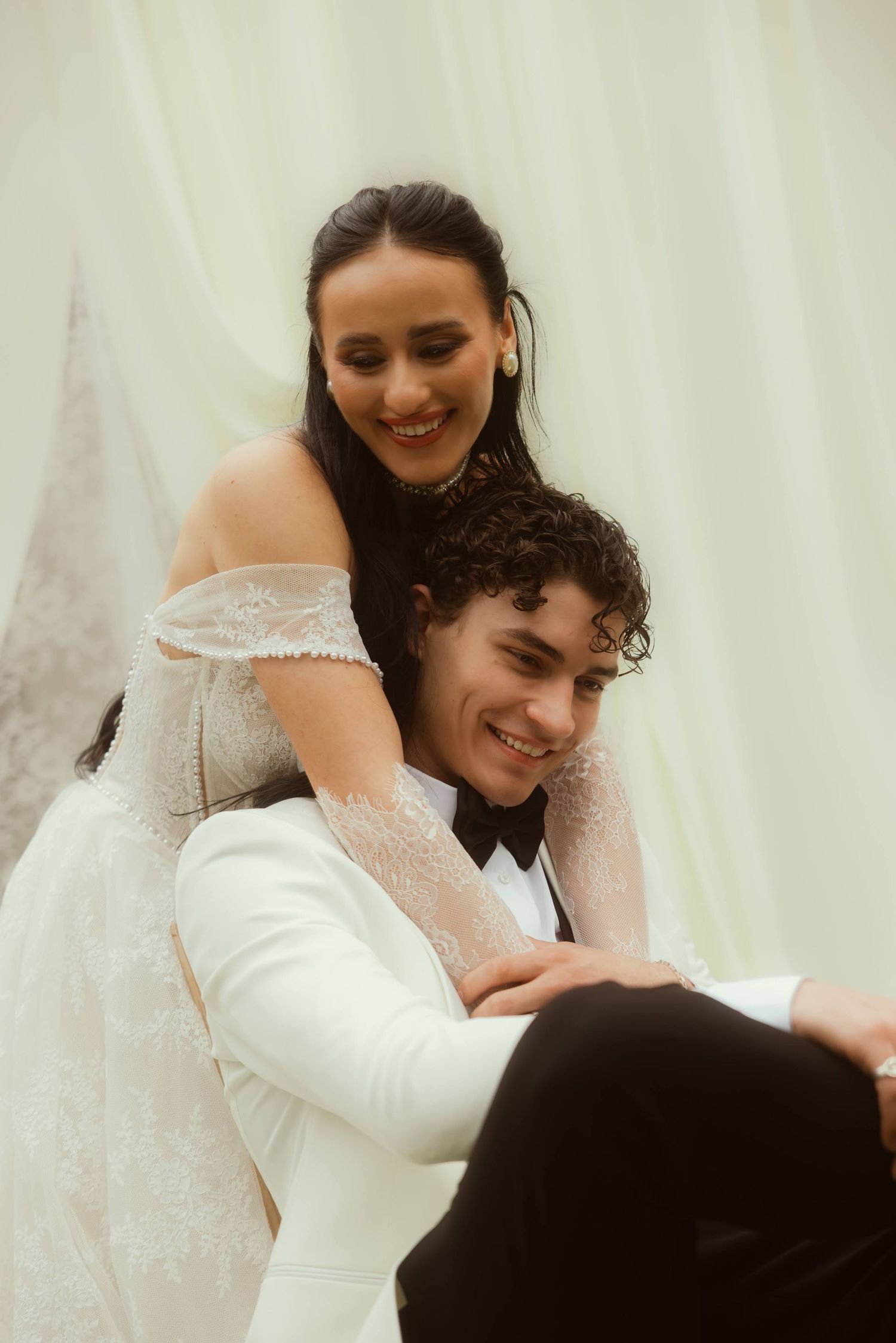 Bride in lace off-shoulder gown embraces smiling groom in white tuxedo jacket, seated together after their Ojai elopement.
