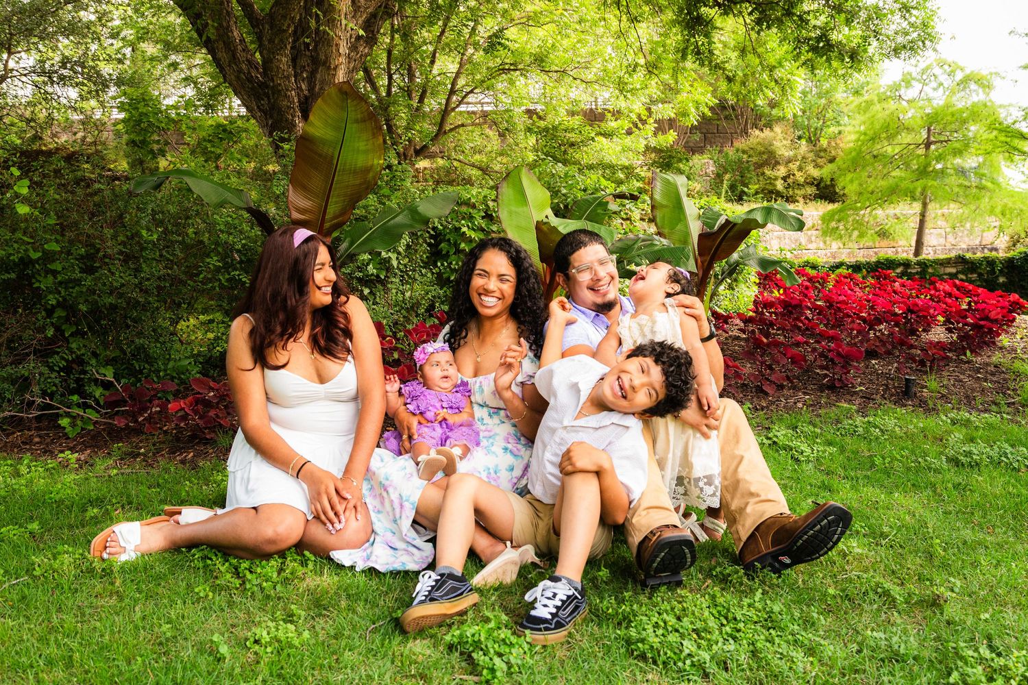 Happy family laughing together on green grass in a garden with red flowers and lush trees.