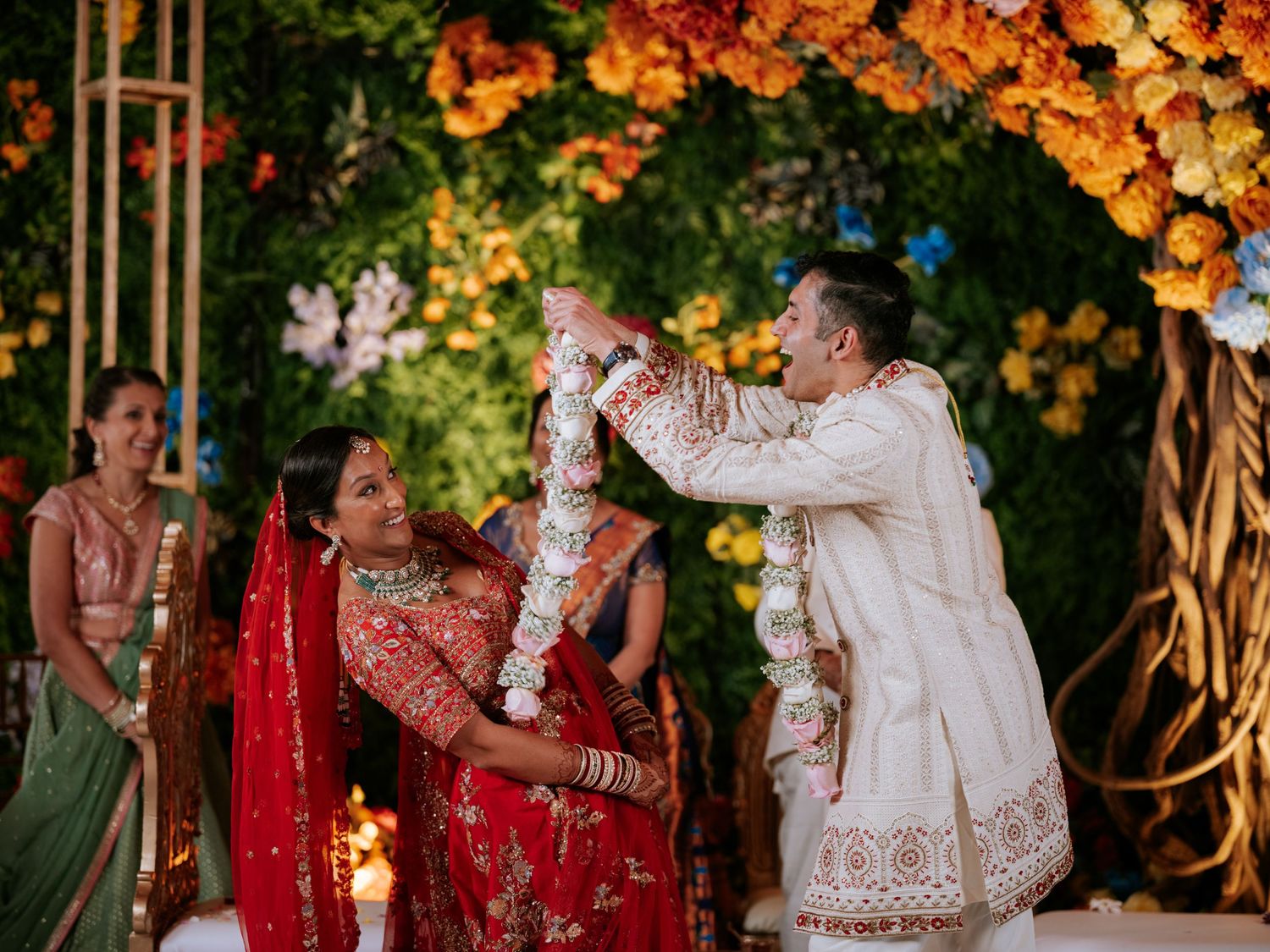 Indian bride and groom exchange garlands during traditional Hindu wedding ceremony with vibrant floral backdrop.