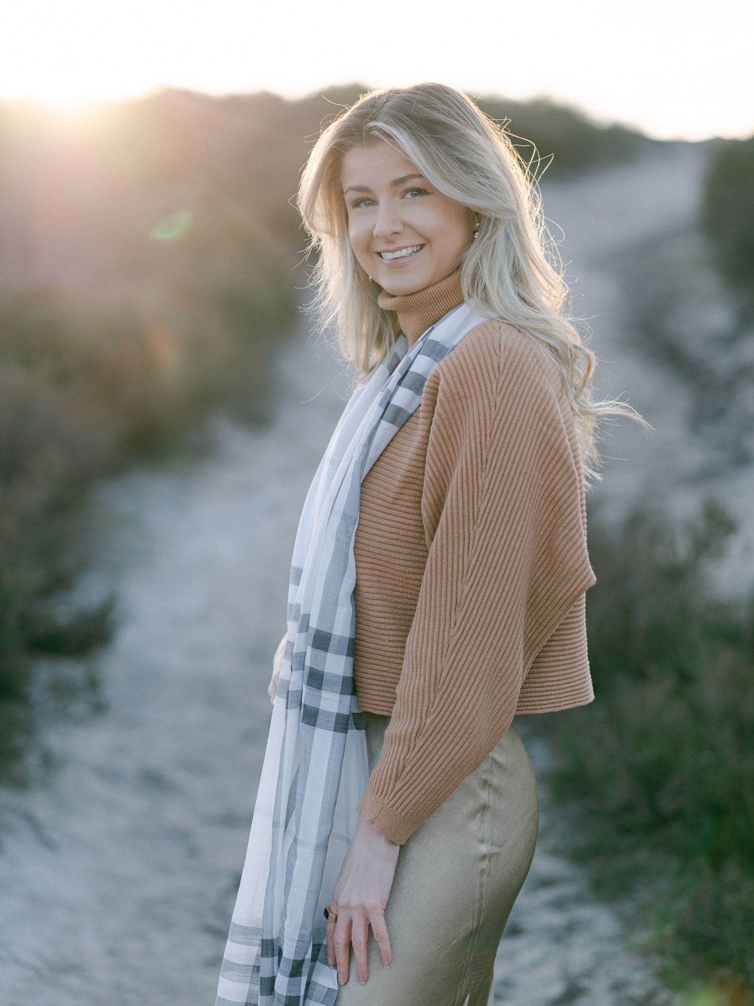 Smiling blonde woman in camel knit sweater and plaid scarf poses on sandy path at golden hour sunset.